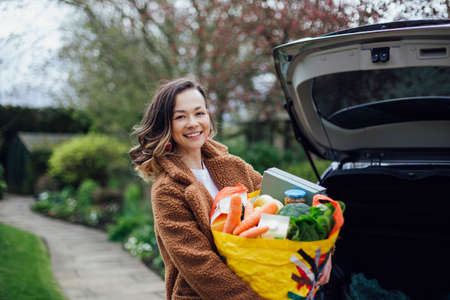 A young woman smiling, looking at the camera and holding a reusable carrier bag filled with groceries. She is unloading the car after food shopping.の写真素材