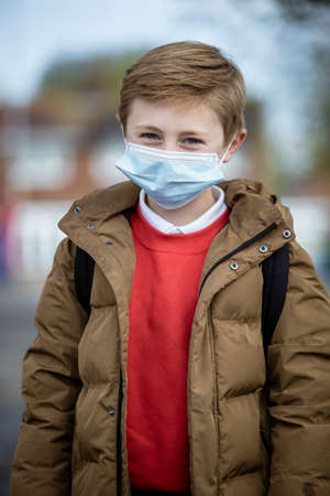 A front-view portrait shot of a young caucasian boy wearing a school uniform, he's smiling and looking at the camera. He is also wearing a protective face mask to help stop the spreadの写真素材