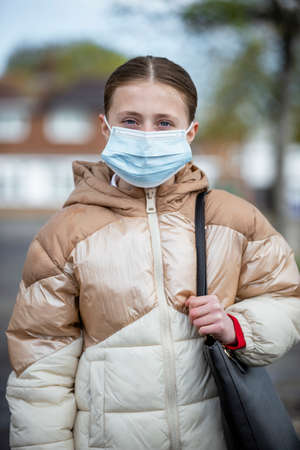 A front-view portrait shot of a young caucasian girl wearing a school uniform and coat, she's smiling and looking at the camera. She is also wearing a protective face mask to help stop the spreadの写真素材