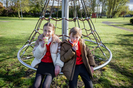 A front view shot of a young caucasian brother and sister after a day at school, they're looking at the camera and smiling while having a rest and enjoying a drink of juice near a public park together.の写真素材