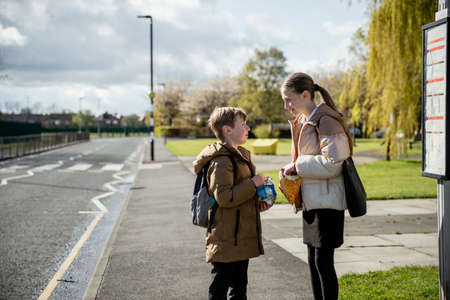 A shot of a young brother and sister standing on a street pathway, they are waiting at a bus stop for a bus home from school, they are enjoying crisp snacks together.の写真素材