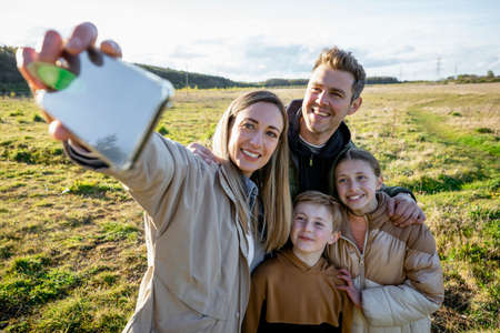 A front-view shot of a caucasian mother and father with their son and daughter, they're all taking a break from walking by smiling for a selfie with a smart phone while wearing casual clothing for a walk outdoors.の写真素材