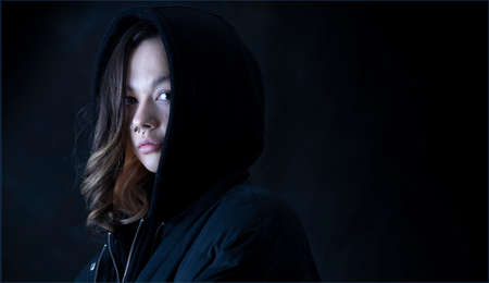 A young woman wearing her hood up, looking at the camera. The room is dark and only her face is lit up.の写真素材
