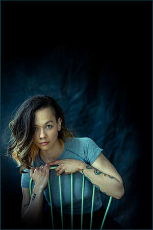 A young contented, trendy woman posing for the camera. She is sitting on a chair and hanging on while leaning to one side and looking at the camera.の写真素材