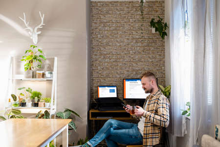 A shot of a home office workspace desk with two monitors, internet banking is displayed on the screens. A man is sitting using his phone by his desk.の写真素材