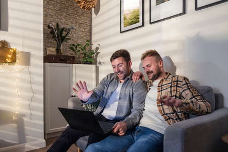An LGBTQI same sex male couple sitting on the sofa in their living room relaxing, they are using a laptop and waving at their friend on a video call.の写真素材