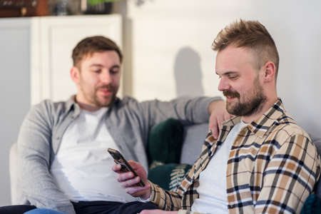 A side-view shot of an LGBTQI same sex male couple sitting in their living room relaxing and using a smart phone.の写真素材