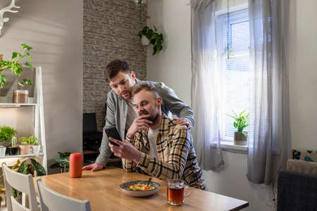 An LGBTQI same sex male couple sitting at a table in their living room relaxing, one man is using his smart phone and he has a plate of pasta.の写真素材
