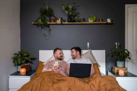 A front-view shot of an LGBTQI same sex male couple lying in their bed in their bedroom relaxing, they are using a laptop and smart phone.の写真素材