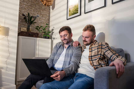 A front-view shot of an LGBTQI same sex male couple sitting on their sofa in their living room relaxing, they are using a laptop.の写真素材