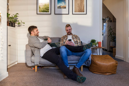 A front-view shot of an LGBTQI same sex male couple sitting in their living room relaxing and using a smart phone.の写真素材