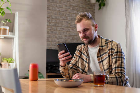 A side-view shot of a young man sitting at a table in his living room relaxing, he is using his smart phone and he has a plate of pasta near by.の写真素材