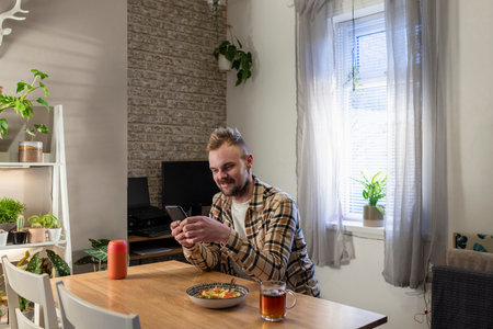 A side-view shot of a young man sitting at a table in his living room relaxing, he is using his smart phone and he has a plate of pasta near by.の写真素材