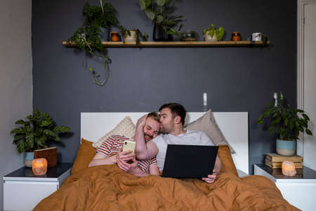 A front-view shot of an LGBTQI same sex male couple lying in their bed in their bedroom relaxing, they are using a laptop and smart phone.の写真素材