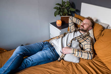 A high angle view shot of a young man lying on his bed in his bedroom relaxing, he has his eyes closed and a hot water bottle by his side.の写真素材