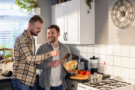 A side-view shot of an LGBTQI same sex male couple standing in their kitchen together, one man is making pasta while the other man talks to him.の写真素材