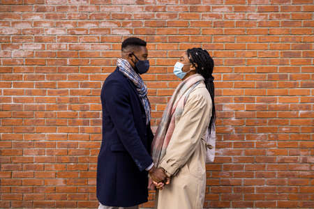 Couple with face coverings standing face to face by brick wall and holding handsの写真素材