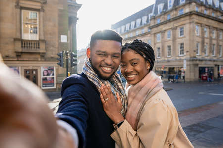 Cheerful couple in town taking selfie with mobile phone, woman with hand on man's chestの写真素材