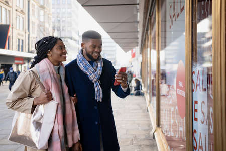 Cheerful couple window shopping and checking mobile phone, smiling and looking through store windowの写真素材