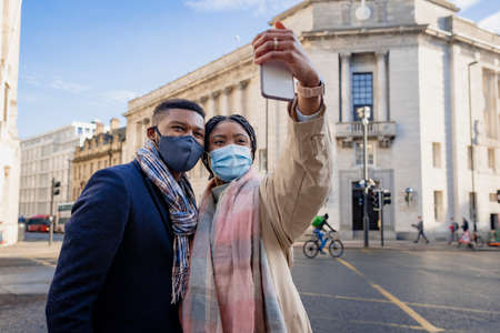 Couple in town wearing face masks holding mobile phone and taking selfieの写真素材