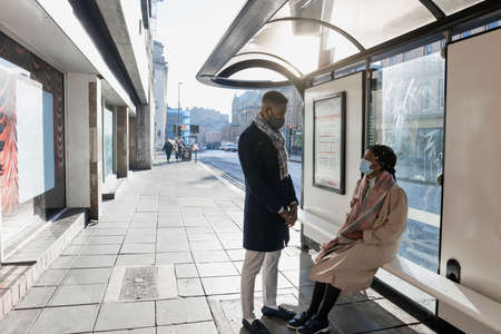 Couple wearing face masks in bust stop, woman sitting on banch, man standing to face herの写真素材