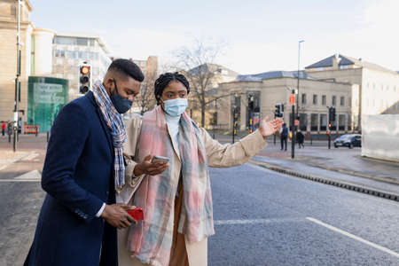 Couple wearing face masks in town waiting for taxi, woman holding mobile with arm outの写真素材