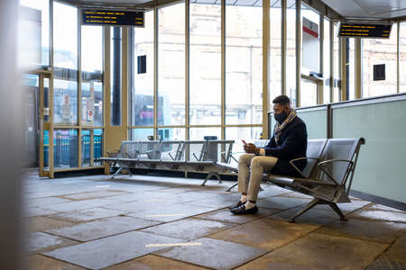 Man wearing face mask sitting on bench in bus terminal using mobileの写真素材