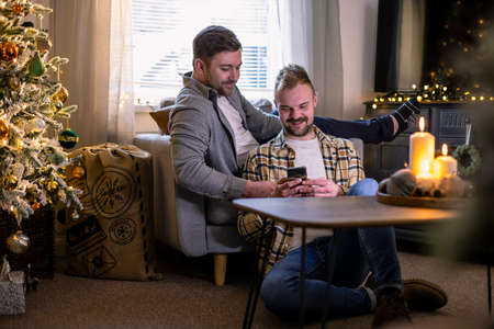 A front-view shot of an LGBTQI same sex male couple sitting in their living room relaxing, one man is showing his partner his smart phone, the house is decorated with christmas decorations.の写真素材