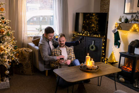 A front-view shot of an LGBTQI same sex male couple sitting in their living room relaxing, one man is showing his partner his smart phone, the house is decorated with christmas decorations.の写真素材