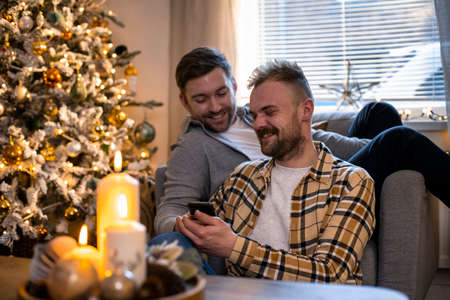 A front-view shot of an LGBTQI same sex male couple sitting in their living room relaxing, one man is showing his partner his smart phone, the house is decorated with christmas decorations.の写真素材
