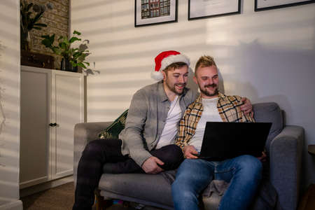 An LGBTQI same sex male couple sitting on the sofa in their living room relaxing, they are using a laptop on a video call. One man is wearing a santa's hat.の写真素材