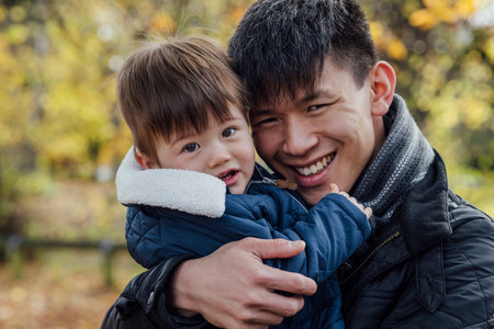 A young man carrying his baby boy in his arms while standing outside and embracing him in nature in Northumberland, North East England. They are both looking at the camera and smiling.の写真素材