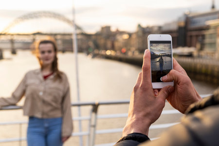 A young couple spending the day in Newcastle Upon Tyne. The woman is posing for a photo her boyfriend is taking on a mobile phone while she stands on a bridge with the River Tyne behind her.の写真素材