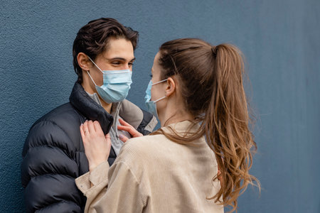 A young couple spending the day in Newcastle Upon Tyne. They are embracing each other while leaning against a wall while smiling and looking at each other. They are wearing protective face masks.の写真素材