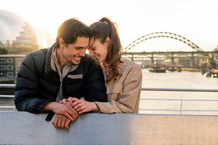 A young couple spending the day in Newcastle Upon Tyne together. They are standing on a bridge with the river tyne and other bridges behind them while embracing each other and looking happy.の写真素材