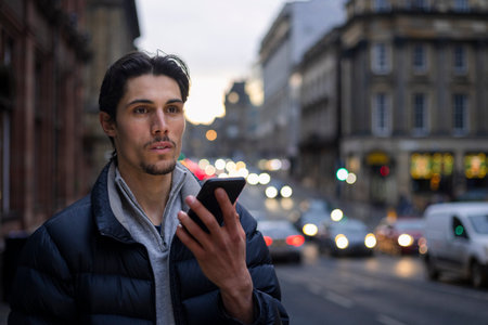 A young man standing on a city street in Newcastle Upon Tyne in the evening while talking to a friend on his mobile phone. He is waiting on a taxi.の写真素材