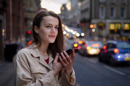 A young woman standing on a city street in Newcastle Upon Tyne in the evening while talking to a friend on her mobile phone. She is waiting on a taxi.の写真素材