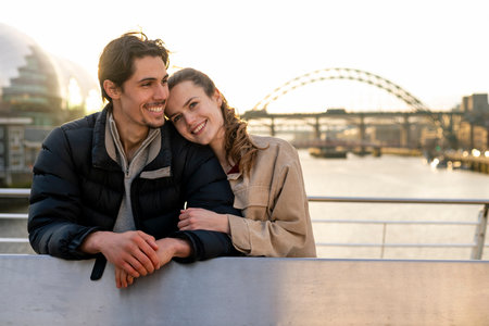 A young couple spending the day in Newcastle Upon Tyne together. They are standing on a bridge with the river tyne and other bridges behind them while embracing each other and looking happy.の写真素材