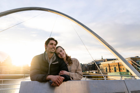 A wide-view shot of a young couple standing side by side looking at the view together on a bridge in the city, they are in love.の写真素材