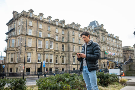 A wide-view shot of a young man standing in the city centre, he is using his smartphone to send a text message.の写真素材