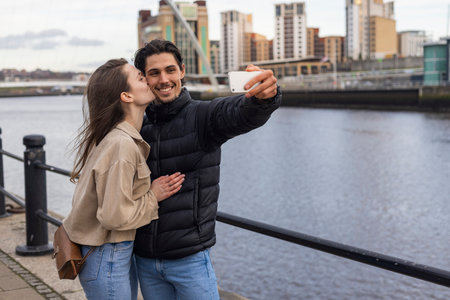 A wide-view shot of a young couple standing together kissing and taking a selfie, they are in love.の写真素材