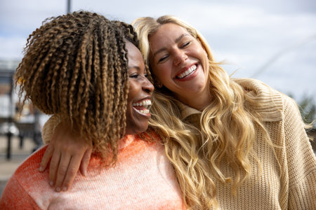 Front-view shot of two mid adult female friends standing in a city street in Newcastle, England. They are wearing casual clothing and sharing a laugh with their heads together.の写真素材