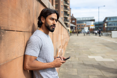 Wide-view shot of a mid adult man standing in a city street, leaning against a wall in Newcastle, England. He is wearing casual clothing while looking away, using a smartphone.の写真素材