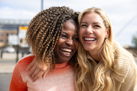 Front-view shot of two mid adult female friends standing in a city street in Newcastle, England. They are wearing casual clothing and sharing a laugh with their heads together.の写真素材