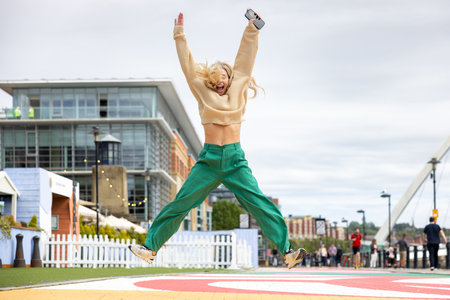 Front-view full-length shot of a mid adult woman jumping with her arms in the air in a city centre street, in Newcastle, England. She is smiling and happy.の写真素材