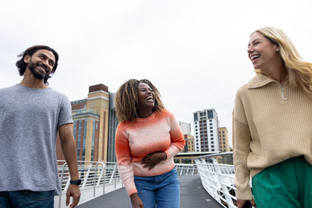 Front-view shot of three mid adult friends crossing a bridge in Newcastle, England. They are wearing casual clothing and walking side by side, smiling and chatting together.の写真素材