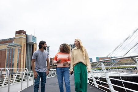 Front-view shot of three mid adult friends crossing a bridge in Newcastle, England. They are wearing casual clothing and walking side by side, smiling and chatting together.の写真素材