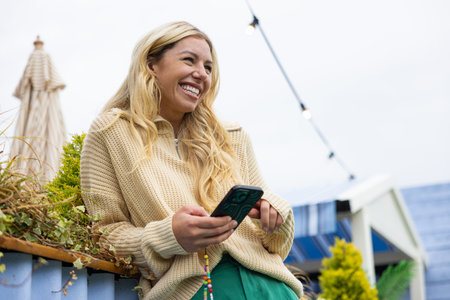 Wide-view shot of a mid adult woman standing in a city centre street, leaning against a wall in Newcastle, England. She is smiling and looking away while using a smartphone.の写真素材