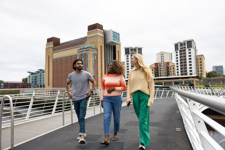 Front-view shot of three mid adult friends crossing a bridge in Newcastle, England. They are wearing casual clothing and walking side by side, smiling and chatting together.の写真素材