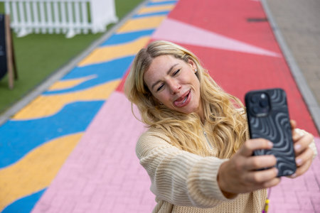 Front-view selfie of a woman standing in a city centre street, in Newcastle, England. She is smiling while taking a selfie on a smartphone, she is sticking out her tongue.の写真素材